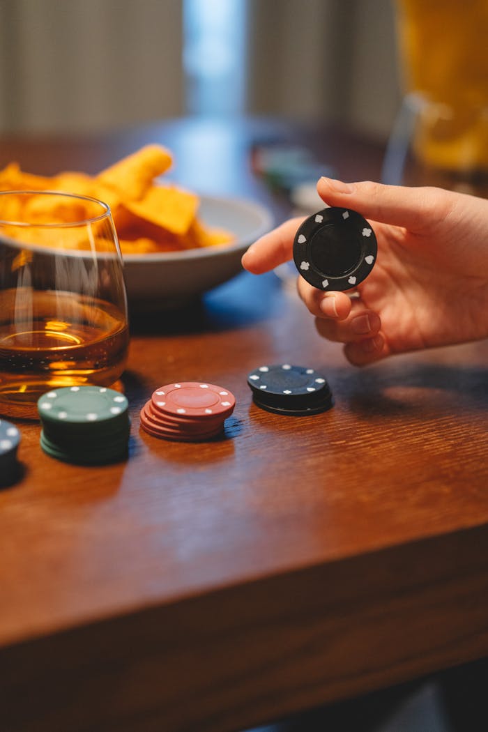 Close-up of a hand holding a poker chip with whiskey and snacks on the table.